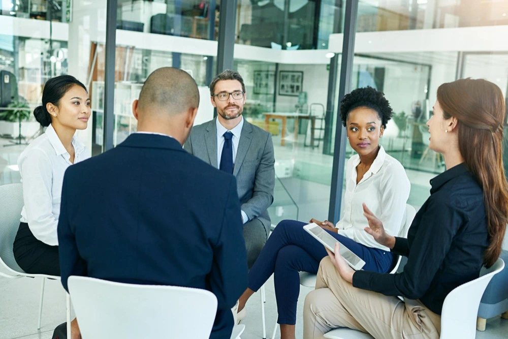 Cinco profissionais participam de uma reunião corporativa, sentados em círculo em um escritório moderno com paredes de vidro, enquanto uma mulher apresenta suas ideias ao grupo.
