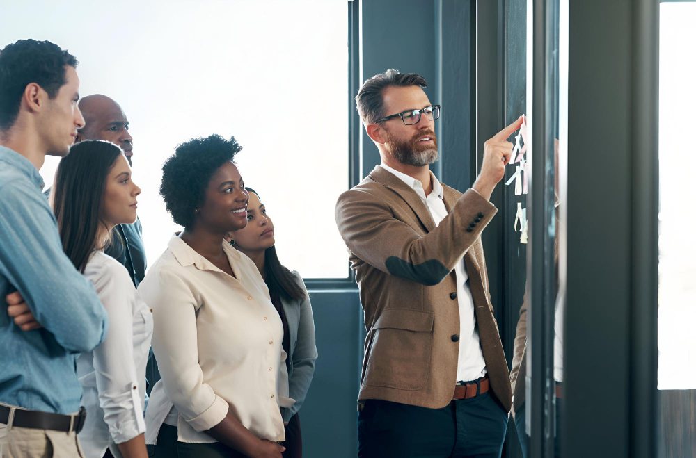 Seis profissionais estão reunidos em um ambiente corporativo, acompanhando uma apresentação de gestão de pessoas por um homem de barba, óculos e blazer marrom. Ele está em pé, apontando para um painel vertical, enquanto os demais participantes observam atentamente as informações apresentadas. O grupo é formado por homens e mulheres com diferentes características físicas, vestidos com roupas sociais e casuais. O escritório possui grandes janelas, que permitem a entrada de luz natural. A imagem representa uma reunião de equipe, destacando a colaboração, a troca de ideias e o planejamento estratégico no ambiente profissional.