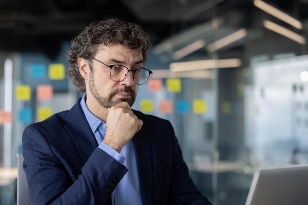 Homem de cabelos castanhos ondulados, barba curta e óculos, vestindo blazer azul-marinho e camisa social azul-clara, aparece sentado em um ambiente corporativo, analisando informações na tela de um notebook. Ele mantém uma das mãos apoiada no queixo e apresenta uma expressão séria e concentrada. Ao fundo, o escritório possui divisórias de vidro, iluminação moderna e notas adesivas coloridas organizadas em uma superfície. A imagem representa um momento de análise e reflexão profissional, destacando a concentração, a tomada de decisões e o planejamento estratégico no ambiente de trabalho.